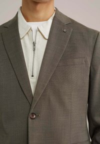 Barna, textúrázott blézer gombos záródással, fehér, cipzáras dzseki felett viselve, amely szőtt mintával és világos gallérral rendelkezik.