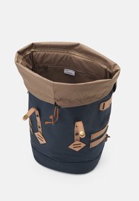 Sac à dos à rabat en tissu bleu marine avec partie supérieure beige. Il comporte des sangles en cuir, des ferrures en laiton et une poche intérieure zippée pour un rangement sécurisé.