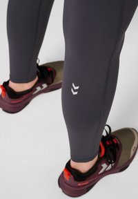 Dopasowane ciemnoszare legginsy o gładkiej fakturze z białym wzorem na łydce, zestawione z burgundowymi butami sportowymi z czarnymi i czerwonymi akcentami.
