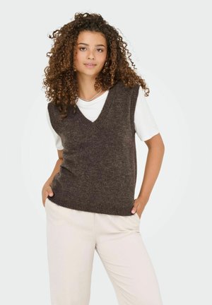 Jonge vrouw met krullend haar die een donkerbruine gebreide vest over een wit shirt en lichtbeige broek draagt, met haar handen in haar zakken, staand.