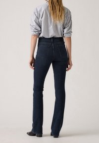 Femme portant un jean bleu foncé et une chemise blanche à manches longues avec des rayures verticales noires, debout de dos devant un fond uni.