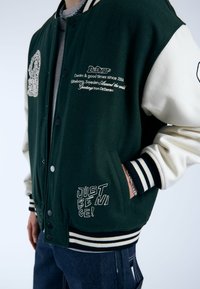 Giacca varsity verde e crema con bottoni neri a pressione. Presenta testo e logo ricamati, polsini a righe e una grande tasca.