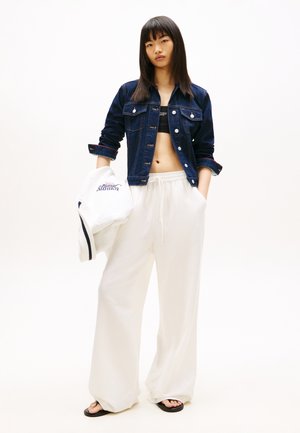 Giovane donna che indossa una giacca di jeans scura, un top corto nero, pantaloni larghi bianchi, sandali neri, con un maglione bianco in mano.