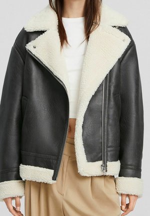 Chaqueta de cuero negra con forro y puños de borrego blanco, llevada sobre una camiseta blanca y pantalones beige de talle alto.