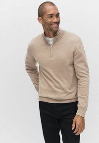 Beżowy sweter zapinany na zamek z dzianiny, z długimi rękawami i ściągaczami, w połączeniu z czarnymi spodniami.