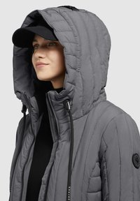 Gray quilted jacket with a large hood, featuring black drawstrings, a black logo patch, and a zippered front. Underneath, a black top can be seen.