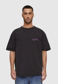 Svart oversized t-skjorte i bomull med rund hals, korte ermer og lila tekstlogo på fronten. Teksturert, lett falmet finish.