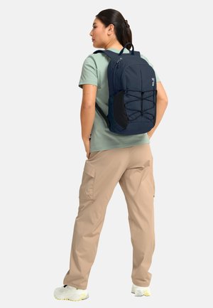 Frau trägt ein salbeigrünes T-Shirt und beigefarbene Cargohosen, trägt einen marineblauen Rucksack mit schwarzen Seitentaschen und einer vorderen Bungee-Schnur.