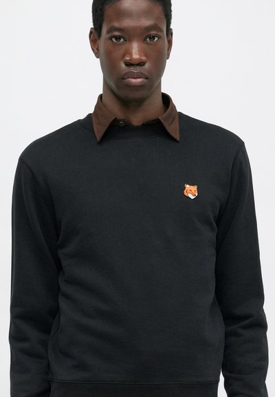 Bărbat îmbrăcat într-un sweatshirt închis la culoare cu un logo în formă de cap de vulpe pe piept și o cămașă cu guler maro dedesubt, stând în fața unui fundal simplu.
