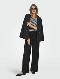 Zwarte dubbelgeknoopte blazer over een gestreept zwart-wit shirt, gecombineerd met zwarte geplooide wijde broek en platte zwarte schoenen.