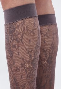 Πόδια που φορούν λεπτές καφέ δαντελωτές καλτσοδέτες με λουλουδάτα και γεωμετρικά σχέδια και ελαστικές ταινίες στην κορυφή, πάνω σε απλό φόντο.