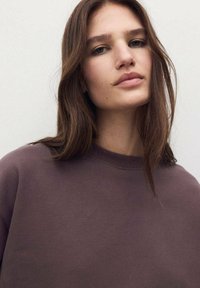 Sudadera marrón con cuello redondo y textura suave, con un corte relajado. El cabello es largo y ondulado, enmarcando el rostro. Fondo minimalista.