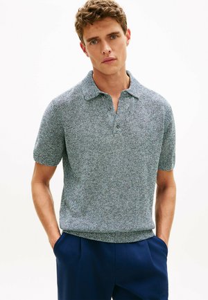 Mężczyzna ubrany w szarą dzianinową koszulkę polo z krótkim rękawem i granatowe spodnie, stoi z rękami w kieszeniach na jednolitym białym tle.