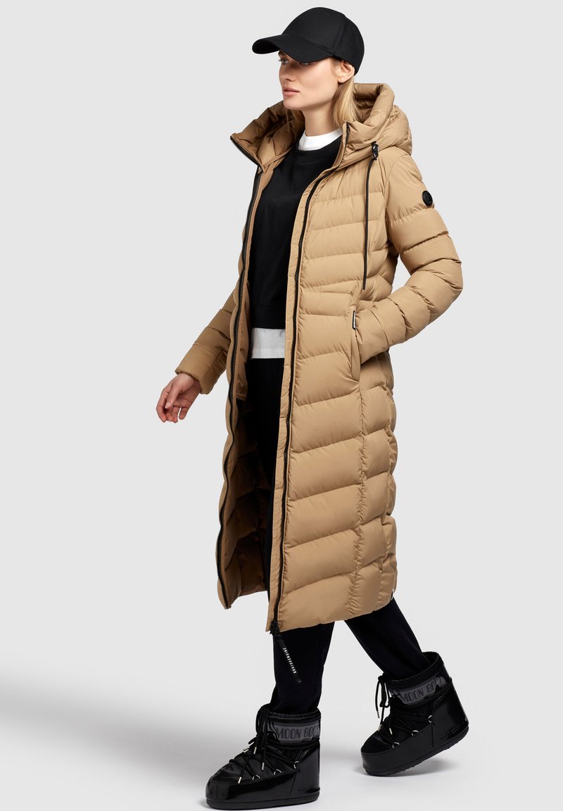 Langer, beigefarbener Puffermantel mit Kapuze, der ein gestepptes Muster, einen Reißverschluss vorne und schwarze Akzente aufweist. Trägt einen schwarzen Pullover und robuste schwarze Stiefel.