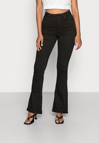 Kvinna som bär högmidjade svarta utsvängda jeans, en vit kort topp och svarta klacksandaler, stående mot en enkel ljus bakgrund.