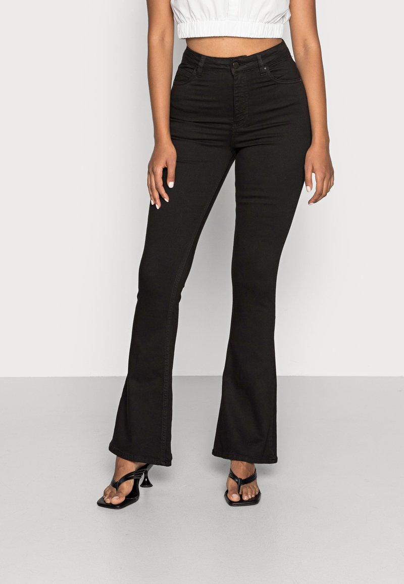 Kvinna som bär högmidjade svarta utsvängda jeans, en vit kort topp och svarta klacksandaler, stående mot en enkel ljus bakgrund.