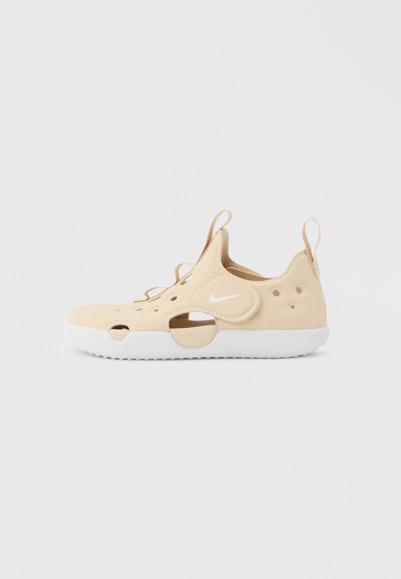Μπεζ αθλητικό παπούτσι χωρίς κορδόνια με λευκή σόλα, διάτρητο επάνω μέρος, ταινίες έλξης στη φτέρνα και τη γλώσσα, και λευκό λογότυπο Nike swoosh στο πλάι.