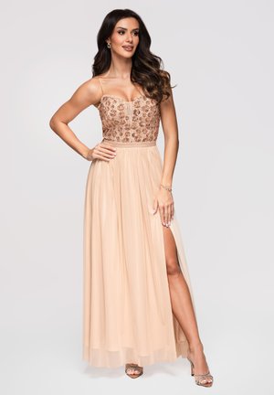 Femme aux longs cheveux foncés portant une robe beige sans manches avec un corsage embellie et une fente haute à la cuisse, posant avec une main sur la hanche.