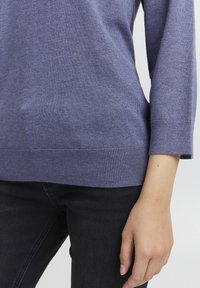 Mīksts, violetas krāsas adīts džemperis ar ribotām malām un trīs ceturtdaļu piedurknēm, apvienots ar melniem džinsiem, uzsverot gludo tekstūru un neformālo dizainu.