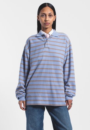 Polo w paski z długim rękawem w jasnoniebieskim kolorze z beżowymi paskami, z klasycznym kołnierzykiem, dwoma guzikami i luźnym krojem. Na zdjęciu niebieskie dżinsy.