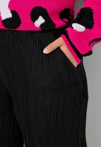 Pantaloni neri plissettati con una texture ondulata, abbinati a un maglione rosa con motivo a fantasia caratterizzato da una stampa leopardata nera e polsini a coste.