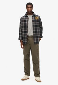 Chemise en flanelle à carreaux noire et grise, avec des boutons rouges et un écusson logo, associée à un pull gris clair et un pantalon cargo vert olive. Baskets beiges.