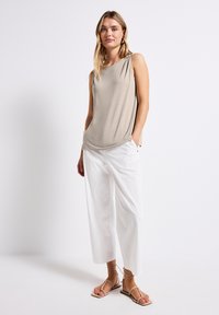 Top beige sin mangas con un ajuste holgado, combinado con pantalones de pierna ancha blancos y sandalias de tiras color marrón, posando frente a un fondo neutro.