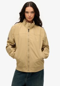 Veste légère beige avec col montant, fermeture à glissière et poches latérales ; possède des poignets élastiques et des coutures discrètes.