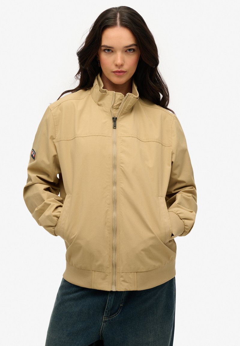 Veste légère beige avec col montant, fermeture à glissière et poches latérales ; possède des poignets élastiques et des coutures discrètes.