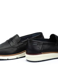Mocasines de cuero negro con un acabado suave, que cuentan con una correa decorativa, suela blanca y granate, y un interior acolchado para mayor comodidad.