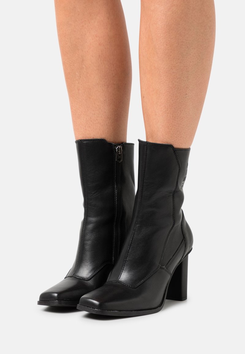 Svarta läderstövlar vid vaden med sidodragkedjor, spetsiga tår och blockklackar, som bärs på bara ben mot en enkel bakgrund.