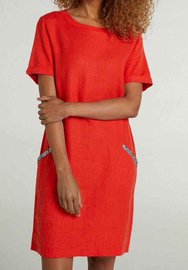Femme portant une robe rouge vif à manches courtes avec des détails scintillants sur les poches latérales, debout, un bras croisé sur l'autre.