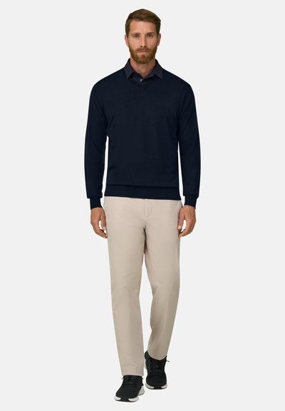 Homme portant une chemise noire à manches longues, un pantalon beige et des baskets noires, debout devant un fond clair uni.
