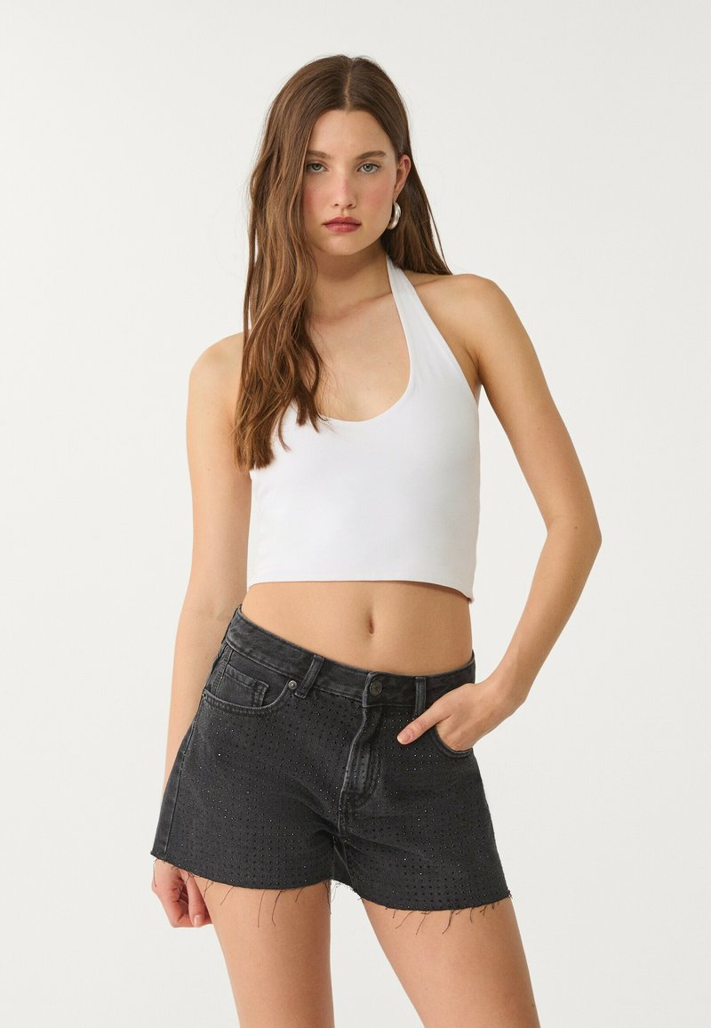 Bijela halter crop majica u kombinaciji s crnim trapericama koje imaju perforirane uzorke i izgrizene rubove. Oba komada imaju prilagođeni dizajn.