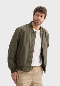 Maslinasto zeleni bomber jakna s uskim krojem, kontrastnim limunasto zelenim patentnim zatvaračem, rebrastim manžetama i izvezenim logotipom na prsima.