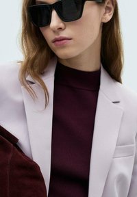 Hellblauer Blazer über einem dunklen burgunderfarbenen Pullover, kombiniert mit übergroßen schwarzen Sonnenbrillen. Glatte Stoffe mit taillierter Passform und klaren Linien.