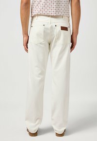 Pantalones de mezclilla blanca con un ajuste relajado, que cuentan con dos bolsillos traseros y un parche de cuero. Textura suave y detalles en las costuras laterales visibles.