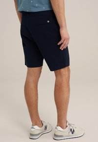 Shorts bleu marine en tissu coton, avec une poche arrière dotée d'un bouton, associés à des baskets beige affichant le logo New Balance.