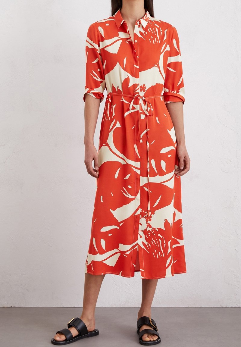 Donna che indossa un vestito midi a maniche lunghe, con bottoni, floreale arancione e crema, con una cintura in vita e sandali neri con fibbia, in piedi davanti a un muro semplice.
