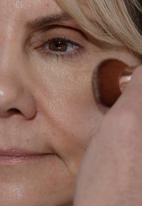 Nahaufnahme eines Gesichts einer Frau mit heller Hautfarbe, feinen Falten und einem Makeup-Pinsel, der Bronzer auf ihrem Wangenknochen aufträgt. Braune Augen und hellblondes Haar.
