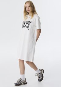 Robe blanche à manches courtes avec une impression graphique noire. Présente une coupe décontractée, arrivant au milieu de la cuisse. Associée à des baskets à motifs et des chaussettes blanches.