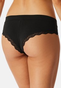 Sous-vêtements hipster pour femmes en dentelle noire sur un corps bronzé, avec un accent sur le dos et le haut des cuisses sur un fond blanc.
