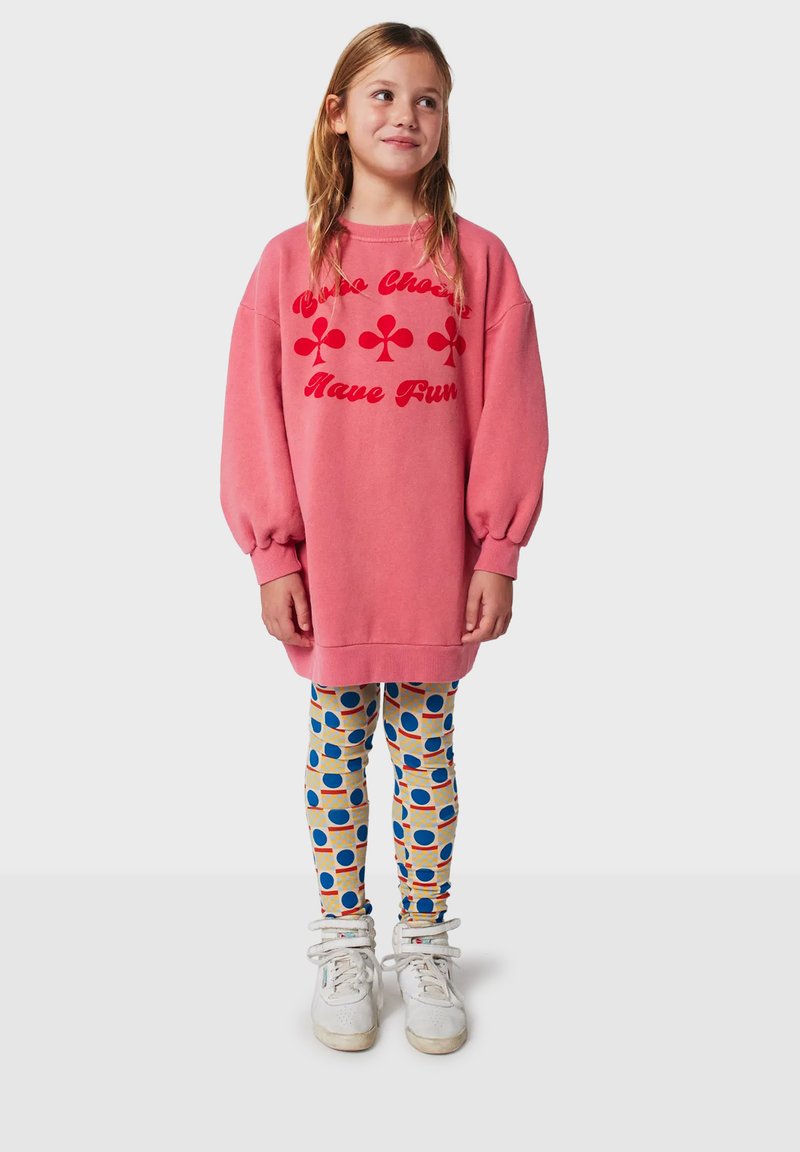 Lyserød oversized sweatshirt med rød tekst og blomster-motiver, kombineret med mønstrede leggings med blå cirkler på cremefarvet baggrund og hvide sneakers.