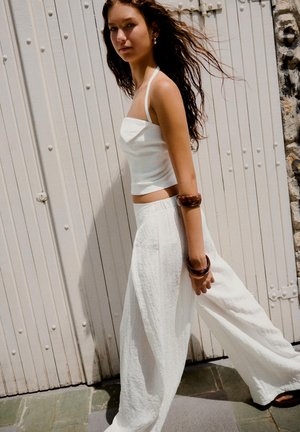 Tânără cu păr lung merge pe lângă o ușă albă de lemn, purtând un top alb cu barete, pantaloni evazați și brățări maro pe brațul stâng.