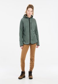 Fleece hoodie in gedempt groen met volledige rits, zwarte accenten en een capuchon. Gepaard met beige broek en bruine wandelschoenen.