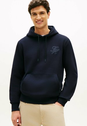 Jonge man die glimlacht, met een marineblauwe hoodie met een voorzak en beige broek, staand met één hand in de zak.