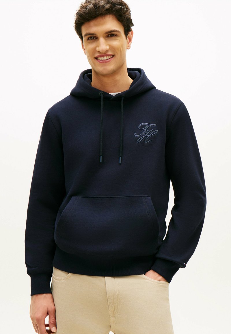 Młody mężczyzna uśmiechający się, ubrany w granatową bluzę z kapturem z przednią kieszenią oraz beżowe spodnie, stojący z jedną ręką w kieszeni.