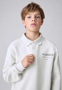 Mladý chlapec se světle hnědými vlasy, který nosí bílou polokošili s dlouhým rukávem s logem "ALLSAINTS", si upravuje jeden knoflík.