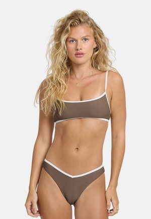 Donna bionda con capelli ondulati che indossa un bikini a righe marroni e bianche con una collana d'oro su uno sfondo chiaro e uniforme.