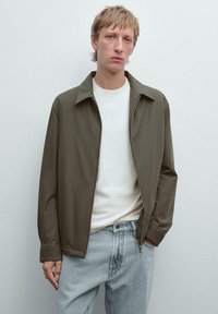 Ung mand iført en olivengrøn zip-up jakke over en hvid skjorte og lyseblå jeans, der står op ad en ensfarvet lys baggrund.